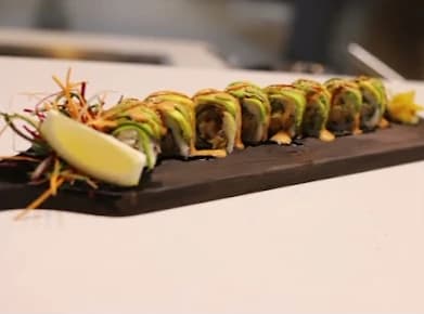 A row of sushi rolls topped with sliced avocado and sauce on a dark wooden board